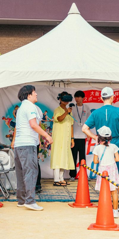 People gather at an outdoor fair booth, enjoying a virtual reality experience in a festive setting.